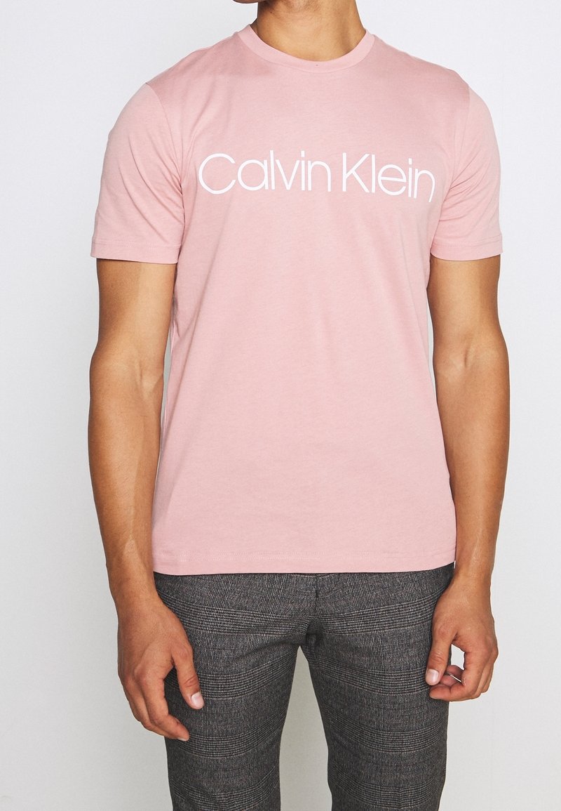 Różowa bawełniana koszulka z logo "Calvin Klein" w kolorze białym, okrągły dekolt, krótkie rękawy, w zestawie z szarymi spodniami w wzory.
