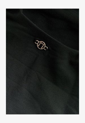 Schwarzer Stoff mit einem subtilen strukturierten Muster, das ein goldfarbenes kreisförmiges Logo-Detail mit einem schwarzen Band verbindet. Netzpaneel oben.