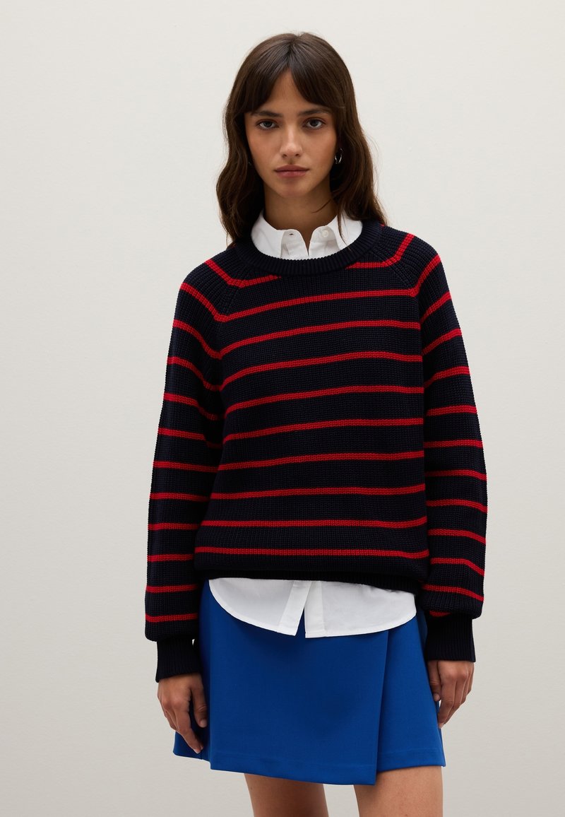 Pull en tricot avec des rayures horizontales bleu marine et rouge, col rond, poignets côtelés, porté sur une chemise blanche à col et une jupe bleue.