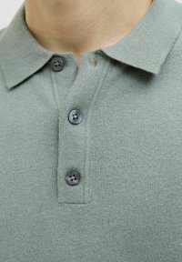 Polo en maille vert clair avec col, présentant trois boutons gris foncé et une texture fine. Aucun motif ni accent visible.