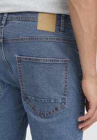 Blugi din denim albastru închis cu finisaj texturat, cu cusături contrastante, un buzunar din spate și o etichetă din piele maro închis la talie.