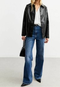 Giacca di pelle nera con colletto, chiusura a bottoni, abbinata a jeans blu a vita alta e a zeppe nere appuntite.
