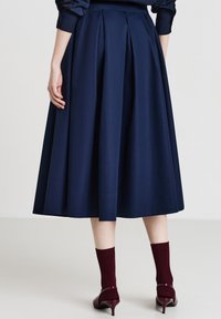 Gonna plissettata blu marino con una texture liscia, lunghezza al ginocchio, dotata di una vita definita e una silhouette fluida, abbinata a calze borgogna e tacchi.