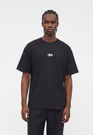 Ung mand med fletninger iført en sort Nike t-shirt med hvidt logo, står foran en ensartet lys baggrund.