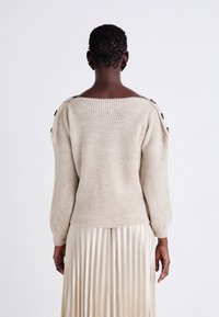 Beež kudumit-sweater, millel on lai kaelus ja nupuvajutused õlgadel, kombineeritud volditud koorevärvi seelikuga. Tekstuuriga kangas.