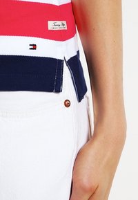 Närbild av en persons midja som bär en randig polo skjorta i röd, vit och marinblå med en Tommy Hilfiger etikett och vita byxor.