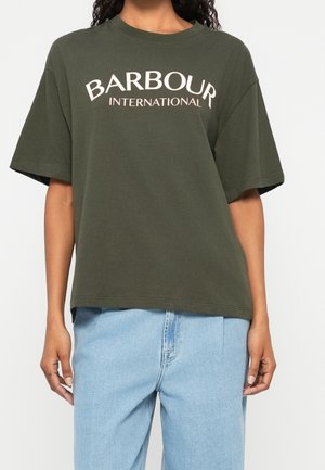 Osoba ubrana w za dużą ciemnozieloną koszulkę Barbour International i jasnoniebieskie dżinsy z wysokim stanem, stojąca na jednolitym tle.