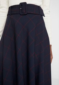 Jupe plissée marine en tissu doux, dotée d'une large ceinture avec une boucle rectangulaire, et d'une coupe évasée avec un motif à carreaux rouges visible.