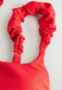 Borsa rossa dal tessuto strutturato con manici arricciati e superficie liscia. La ferramenta comprende un fermaglio dorato per la regolazione della tracolla.