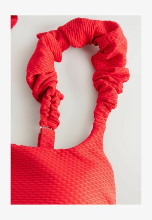 Pöttyös textúrájú piros táska, amely ráncolt pántokkal és sima felülettel rendelkezik. A kiegészítők között egy arany színű klipsz található a pántok állításához.