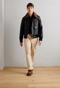 Black leather bomber jacket with a brown fur collar, paired with beige trousers and brown boots, on a model walking on a wooden floor.
