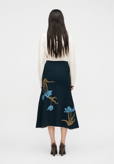 Γυναίκα με μακριά σκούρα μαλλιά, που φοράει λευκό πουλόβερ, μπλε φούστα με μπλε και καφέ ανθικά κεντήματα και καφέ παπούτσια με τακούνια, με την πλάτη στο θεατή.