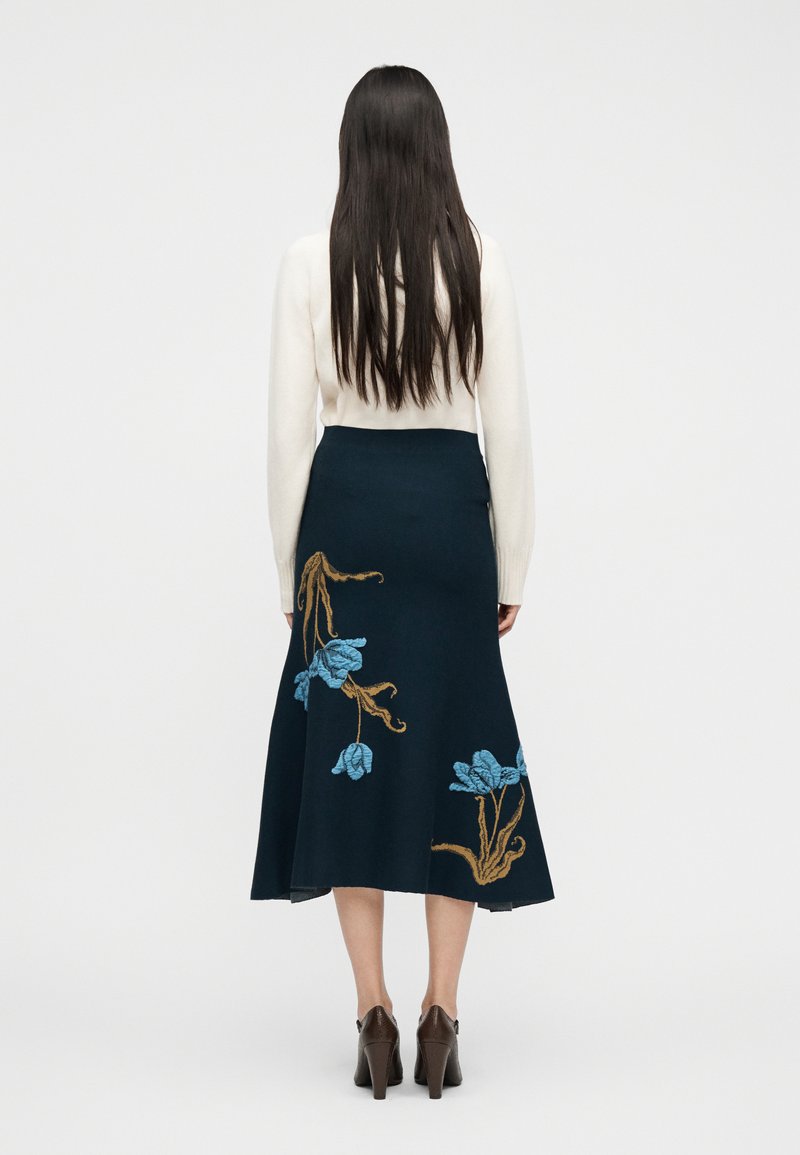 Vrouw met lang donker haar, gekleed in een witte trui, een marineblauwe rok met blauw-bruine bloemrijke borduursels en bruine schoenen met hakken, van achteren gezien.