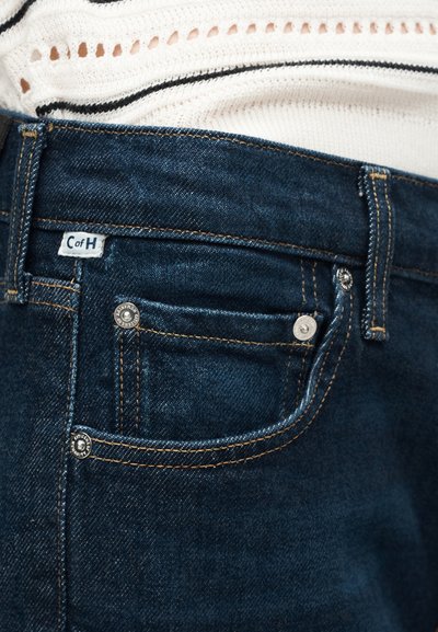 Κοντινό πλάνο τσέπης σκούρων μπλε τζιν με πορτοκαλί ραφές και λευκή ετικέτα "C of H", φορεμένη με λευκό πλεκτό κορυφαίο με μαύρες ρίγες.