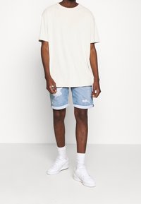 Camiseta de cuello redondo beige, pantalones cortos de mezclilla desgastados en azul claro con dobladillos enrollados, zapatillas blancas y calcetines blancos. Atuendo casual.