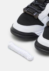 Black mesh and white leather sneakers with a rubber outsole and distinctive stitching, accompanied by a rolled white shoelace.