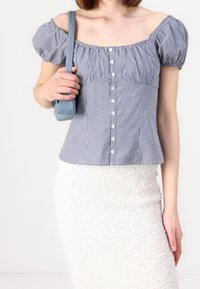 Top a maniche puff in gingham blu e bianco con vita aderente, chiusura frontale con bottoni e collo elastico, abbinato a una gonna bianca con texture.