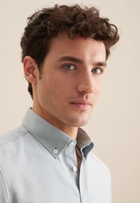 Camicia azzurro chiaro con colletto, con un motivo testurizzato, abbottonata, presenta un colletto interno con motivo a contrasto. Il modello ha i capelli ricci.