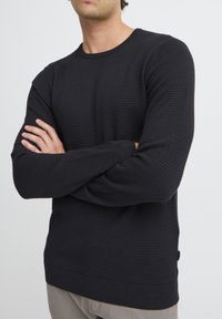 Pulover negru texturat cu un design tip țesătură waffles, mâneci lungi și manșete și tiv ribbotate. Prezentă branding minimal pe partea inferioară.
