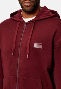 Sudadera con cremallera en color burdeos, hecha de una tela suave, con capucha, cordón con puntas metálicas y un logo brillante de "HUGO" en el pecho.