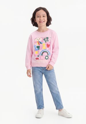 Smilende barn med kort brunt hår iført en lyserød sweatshirt med farverige motiver, blå jeans og hvide sneakers, står mod en hvid baggrund.
