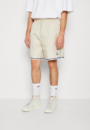 Homme portant un short de basketball beige avec une bordure noire, des chaussettes blanches Nike et des baskets montantes Nike gris clair, debout devant un fond blanc.