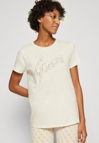 Camiseta de manga corta color crema con un logo "Guess" texturizado y parecido a encaje en la parte frontal; corte holgado y cuello redondo.