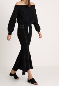 Sudadera negra con escote off-the-shoulder, de corte holgado, con dobladillo acanalado. Combinada con jeans negros de campana con costuras blancas en contraste y mules negras de tacón.