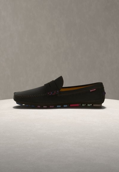 Mocassin en cuir noir avec dessus cousu et semelle en caoutchouc multicolore à texture grainée, présenté sur une surface claire devant un fond gris.