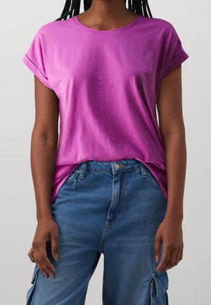 Frau trägt ein leuchtend violettes T-Shirt mit kurzen Ärmeln, das in hochtaillierte blaue Denim-Cargohosen mit aufgekrempelten Ärmel gesteckt ist.