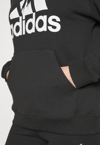 Haut en coton noir avec le logo Adidas blanc. Comprend une poche kangourou et des poignets côtelés. Texture douce et coupe décontractée.