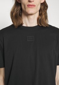 T-shirt noir à manches courtes avec un col rond et une patte texturée noire comportant le mot "HUGO". Tissu lisse avec une coupe ajustée.