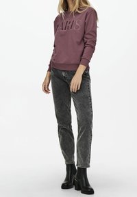 Felpa viola con scritta "PARIS", abbinata a jeans neri sbiaditi e stivaletti neri alla caviglia. Il materiale è morbido con polsini a costine sulle maniche.