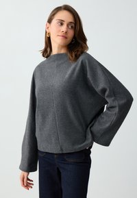 Femme portant des boucles d'oreilles en anneaux dorés, un haut gris ample à manches longues et un jean foncé, debout une main sur la hanche, regardant sur le côté.
