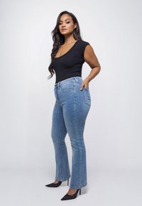 Vrouw met lang zwart haar, gekleed in een zwart mouwloos topje, lichtblauwe jeans en zwarte hoge hakken, staand voor een witte achtergrond.