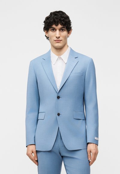 Άνδρας με σγουρά σκούρα μαλλιά που φοράει ανοιχτό μπλε σακάκι και παντελόνι, πάνω από λευκό πουκάμισο με κουμπιά, στέκεται σε απλό φόντο.