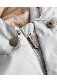 Lys grå jakke med en myk beige foring, med glidelås, messing trykknapper og en teksturert etikett. Nærbilde av stoffet og detaljene.