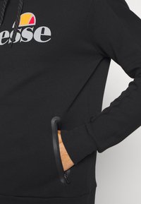 Sweat à capuche noir avec une main dans une poche latérale zippée et un logo partiellement visible en blanc et orange sur la poitrine.