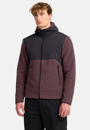 Junger Mann trägt eine zweifarbige braun-schwarze Kapuzenfleecejacke mit Frontreißverschluss und Reißverschlusstaschen an den Seiten, steht vor einem schlichten Hintergrund.