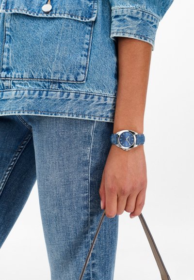 Blue watch with a denim textured strap, silver hardware, and a round face with blue dial. Worn on a hand holding a bag; paired with denim jacket and jeans.