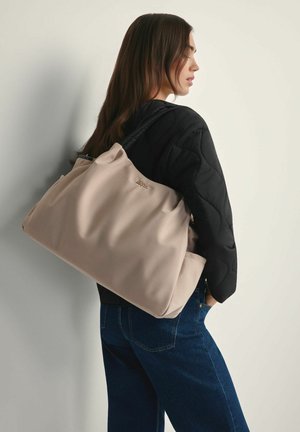 Kobieta w czarnej pikowanej kurtce i niebieskich dżinsach, niosąca dużą beżową torbę na ramię z czarnymi uchwytami, stojąca na tle jasnej ściany.