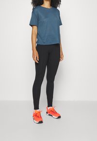 Koszulka sportowa z krótkim rękawem w odcieniu teal, czarne legginsy i jaskrawo pomarańczowe buty do biegania. Koszulka ma teksturowane wykończenie i luźny krój.