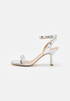 Sandale à talon haut argentée avec texture reptile, bride ouverte sur le devant et bride de cheville avec boucle sur fond blanc.