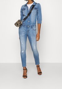 Džínový overal v světle modré barvě, s dlouhými rukávy, zapínáním na knoflíky vpředu a roztržen�ými detaily na kolenou. Doplňky zahrnují malou kabelku přes rameno s leopardím vzorem.