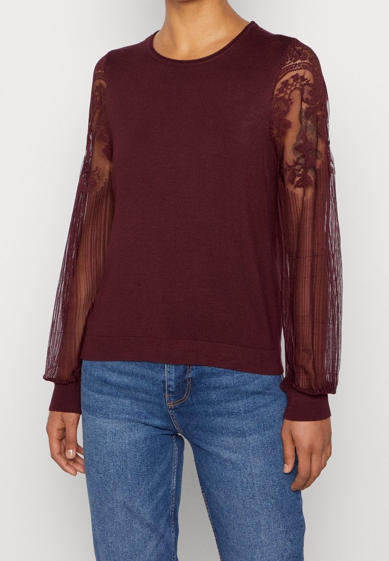 Femme portant un pull bordeaux avec des manches longues en dentelle florale transparente et un jean bleu, se tenant devant un fond uni.