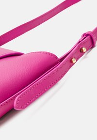 Cinghia di pelle rosa brillante con borchie regolabili in tono oro su uno sfondo bianco.