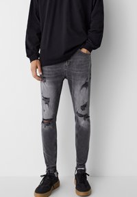 Faded black skinny jeans with distressed details and ripped knees, paired with black sneakers, worn with a loose-fitting black long sleeve shirt.