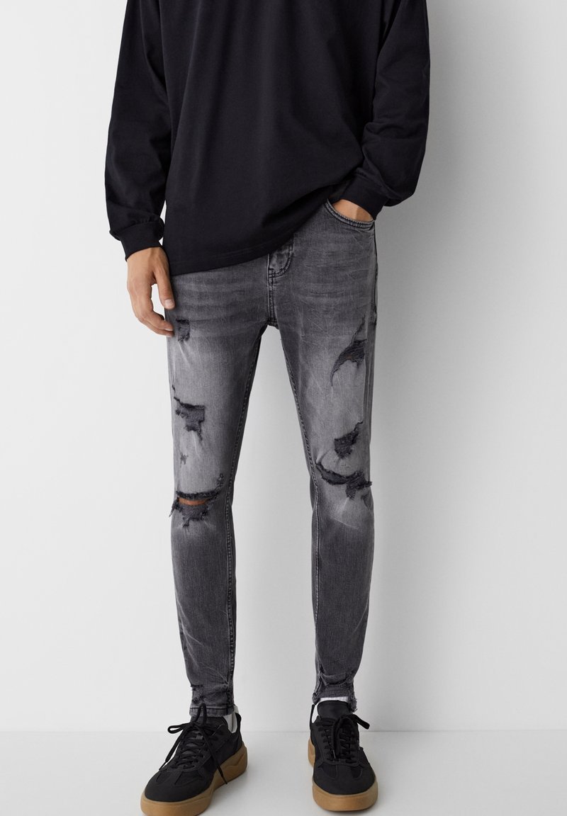 Faded black skinny jeans with distressed details and ripped knees, paired with black sneakers, worn with a loose-fitting black long sleeve shirt.