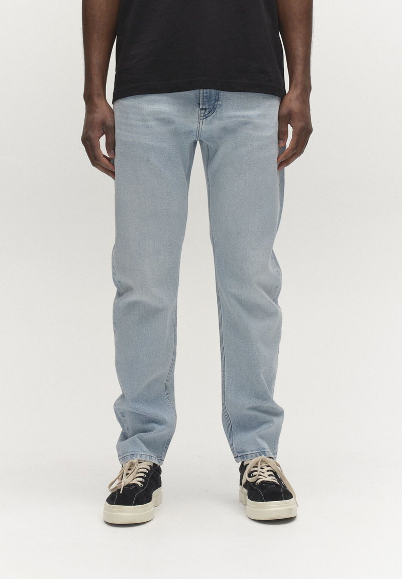 Személy világoskék farmert és fekete, fehér talpú és fűzős tornacipőt visel, tiszta fehér háttér előtt áll.