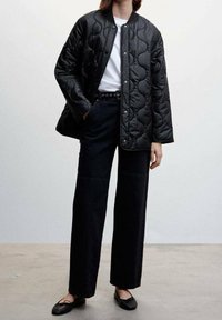 Osoba ubrana w czarną pikowaną kurtkę na białą koszulę, wysoko wypuszczone czarne spodnie z szerokimi nogawkami, czarny pasek i czarne baletki.